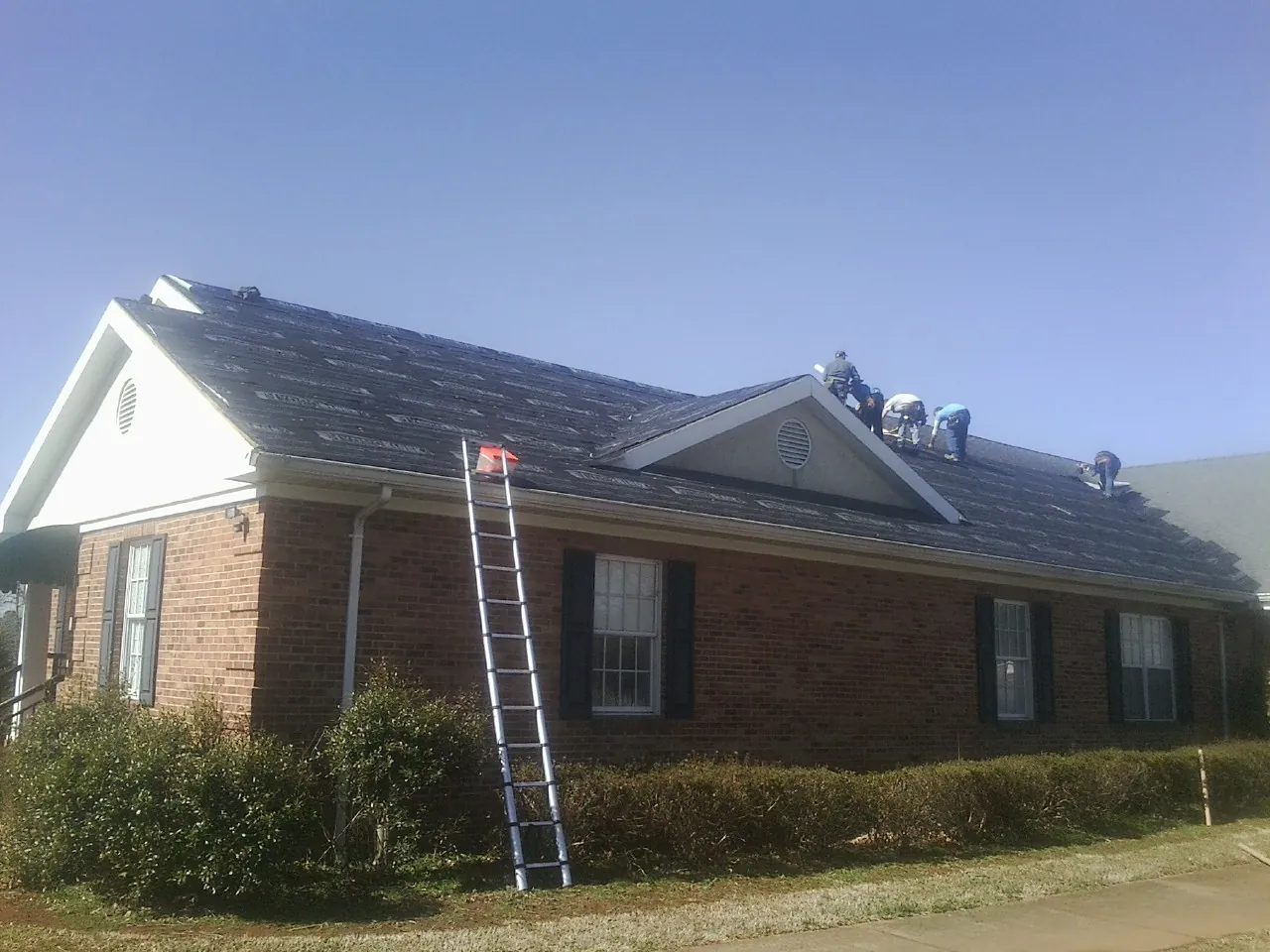 Crew tearing shingles off a brick ranch roof with a ladder set — Metal Roof Repair work by our crew in Shambaugh