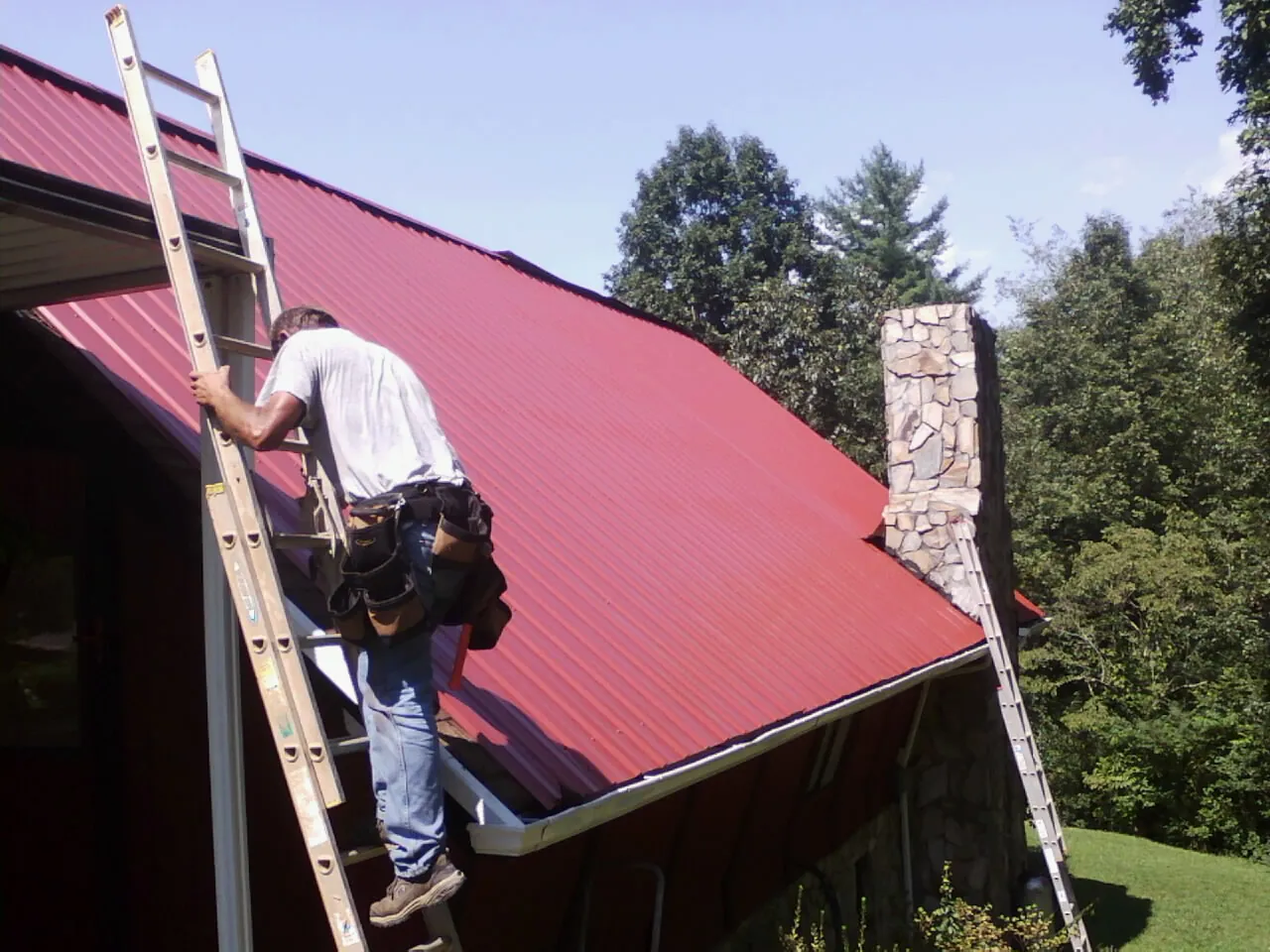 Roofer on a ladder beside a red metal roof and stone chimney — Soffit & Fascia Repair by Roofing Brothers in Shambaugh